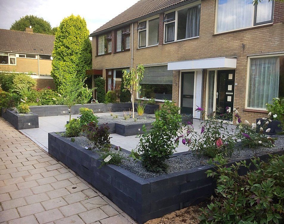 Buitenkant van een huis met een grijs betegeld terras en verhoogde plantenbakken gevuld met planten en grind.