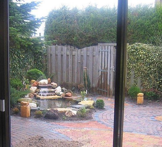 Uitzicht op de achtertuin door een raam: waterpartij, stenen terras, houten schutting en groen.
