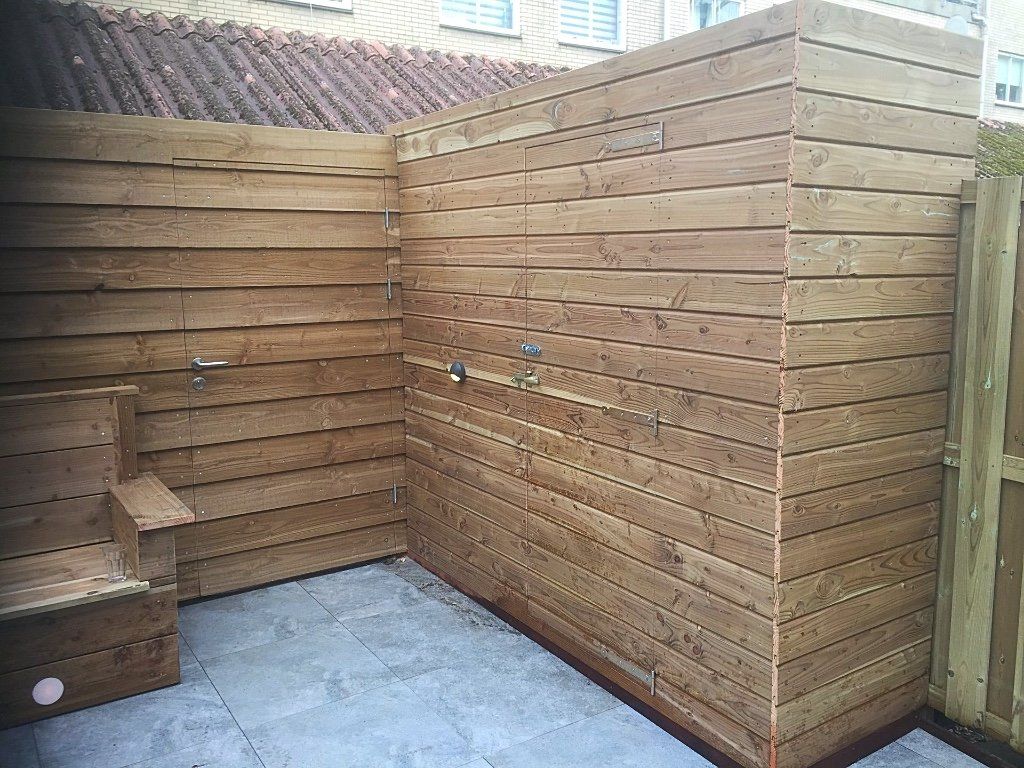 Houten buitendouche met zitje en kranen, geplaatst tegen een muur en schutting op een betegeld terras.