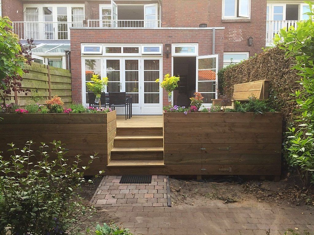 Achtertuin met een houten terras en bloembakken, die leidt naar een bakstenen huis met openslaande deuren en ramen.