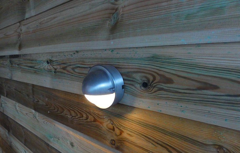 Een ronde, zilverkleurige buitenlamp verlicht een houten muur.