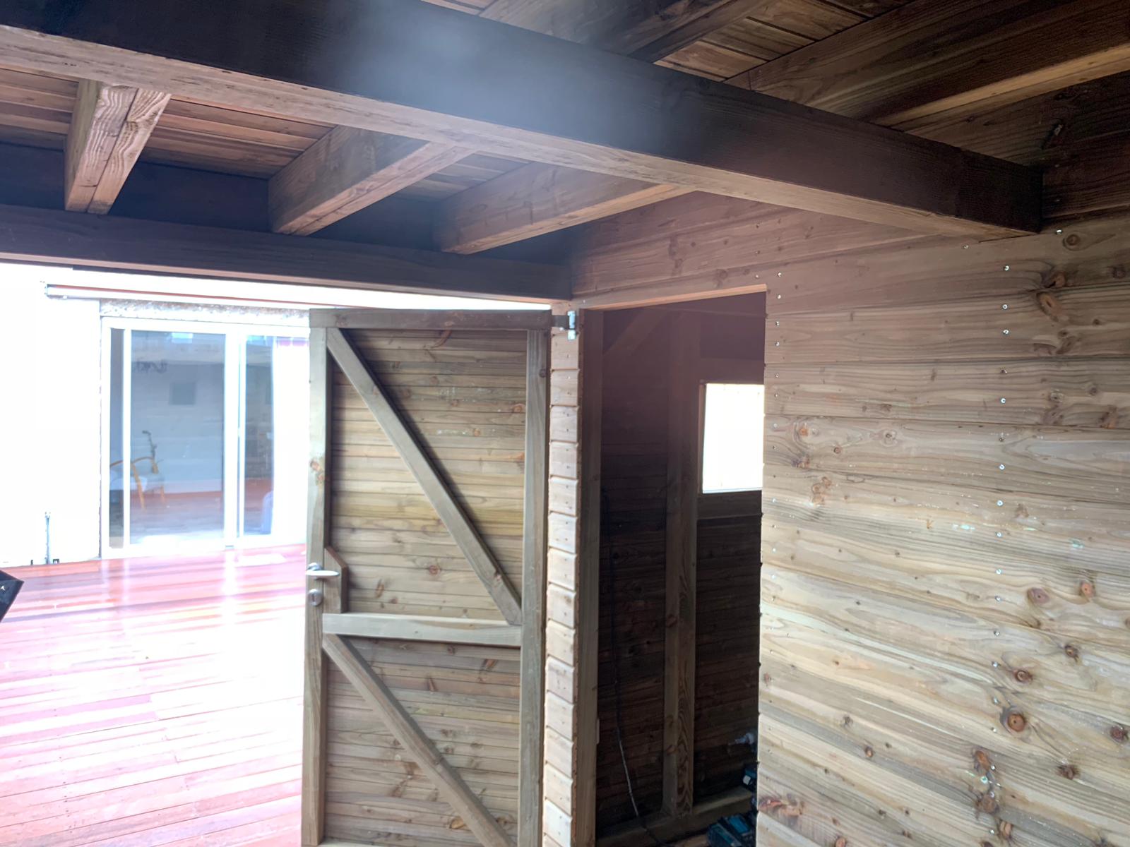 Houten schuurinterieur met open deur, waardoor de veranda en de buitenconstructie zichtbaar zijn.