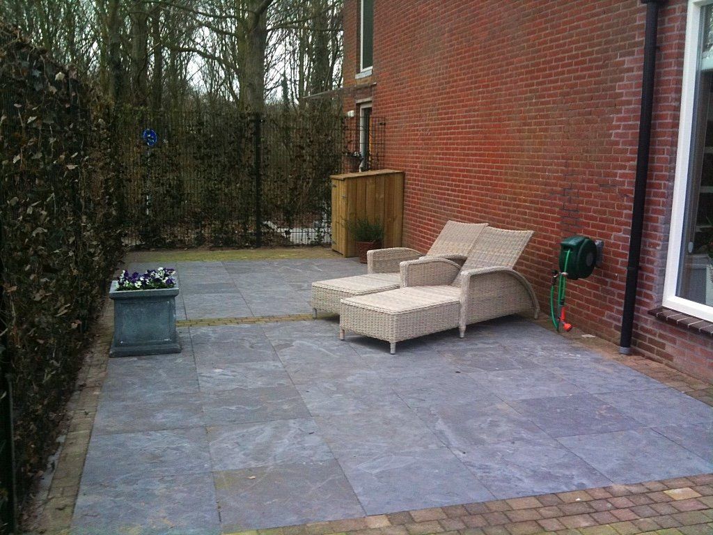 Stenen terras met rieten loungestoelen naast een bakstenen gebouw en een heg.
