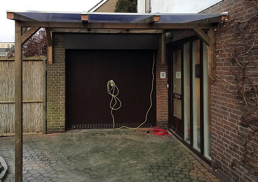 Carport met bruin houten frame, aangebouwd aan een bakstenen gebouw. ​​Donkere garagedeur. Doorzichtig plastic dak.