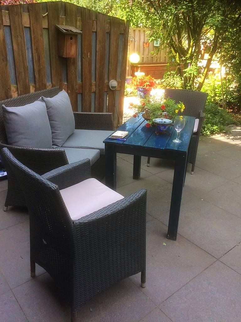 Terrasmeubelset: rieten bank en stoelen met kussens, tafel met planten, houten schutting op de achtergrond.