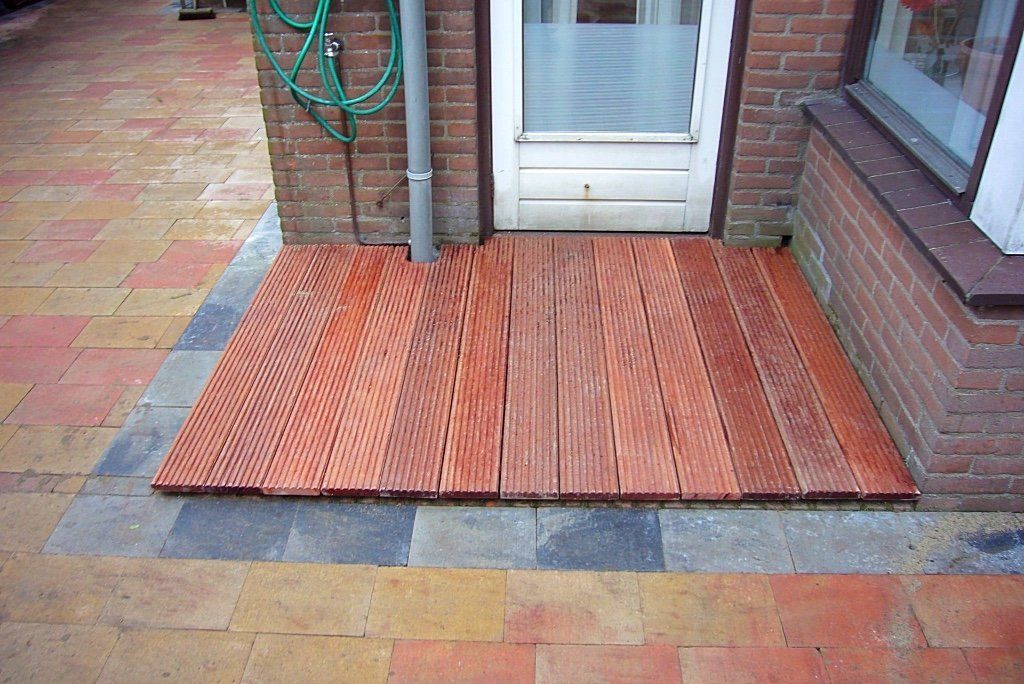 Houten terras bij de voordeur, roodbruine planken, omzoomd door grijze en baksteenkleurige bestrating.