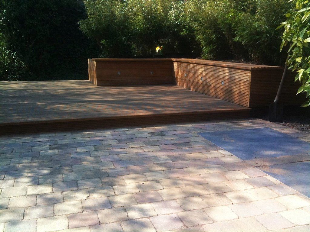 Houten terras met ingebouwde bank in een tuinachtige omgeving, grenzend aan een bestraat terras.