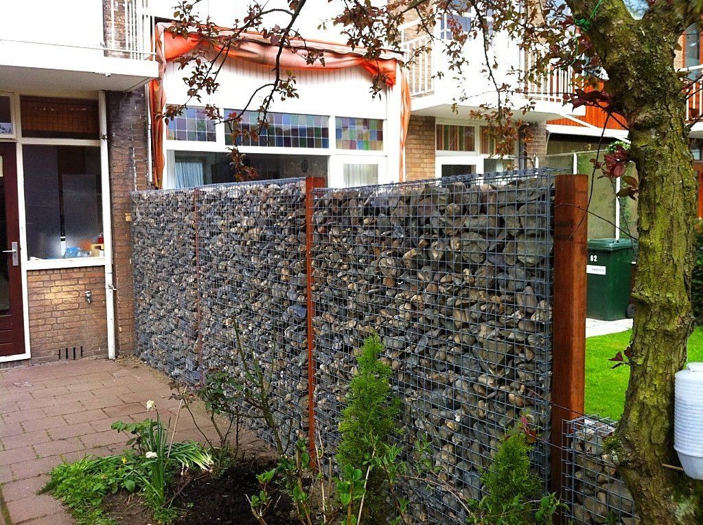 Een schanskorfmuur gevuld met grijze stenen in een woonerf. Er staat een kleine groene struik voor.