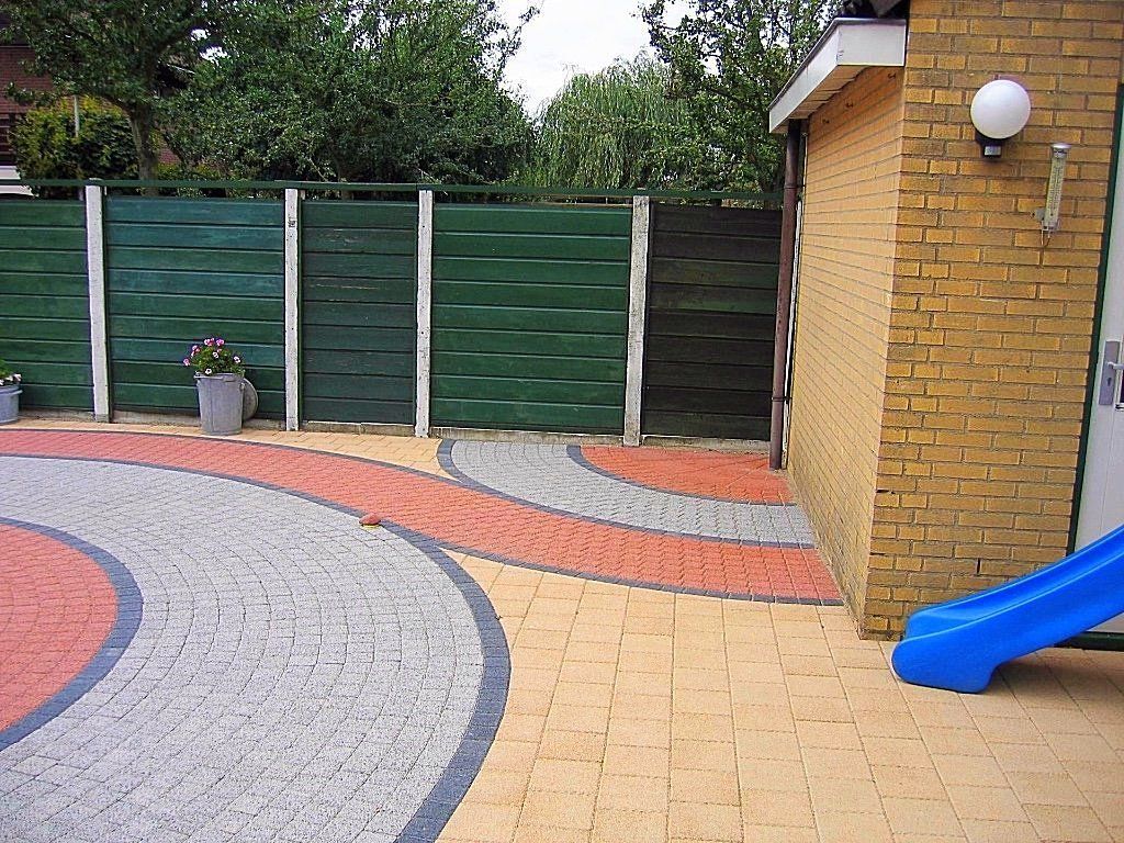Bakstenen terras met blauwe glijbaan, groene schutting en gebogen grijze en rode bakstenen accenten.