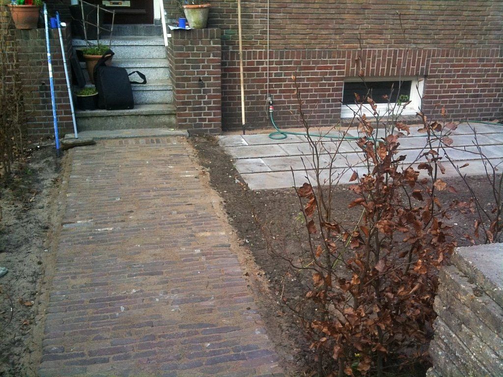 Een bakstenen pad leidt naar een trap, omzoomd door aarde en een bakstenen muur met een klein raam.