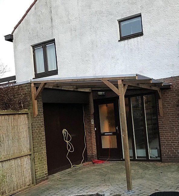 Buitenkant van een huis met een carport boven de voordeur. Bruine garagedeur, bakstenen muur, witte gevel van het huis.