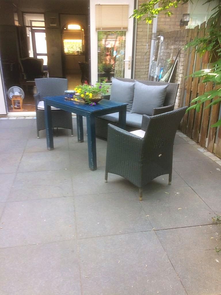 Terras met grijs meubilair: een tafel, een tweezitsbank en twee stoelen. Bakstenen muur, houten schutting en open deur.