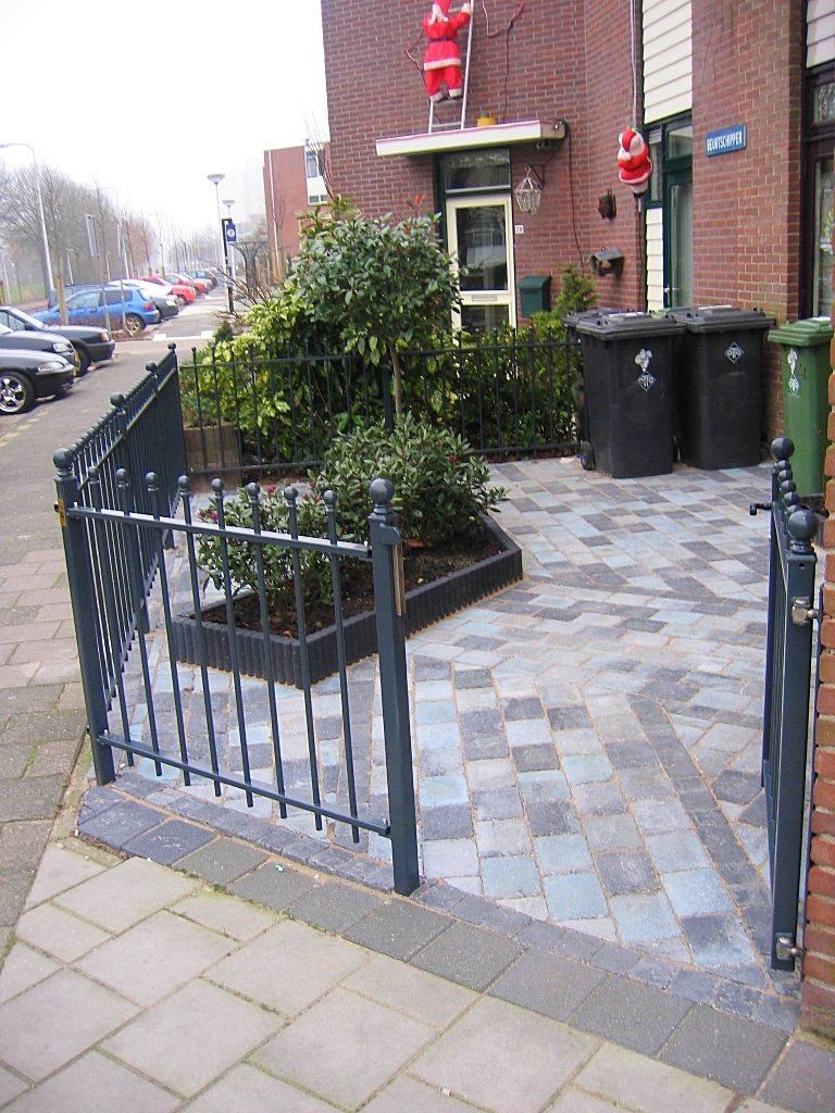 Een stenen terras met een metalen hek, struiken, vuilnisbakken en een huis met kerstversiering.