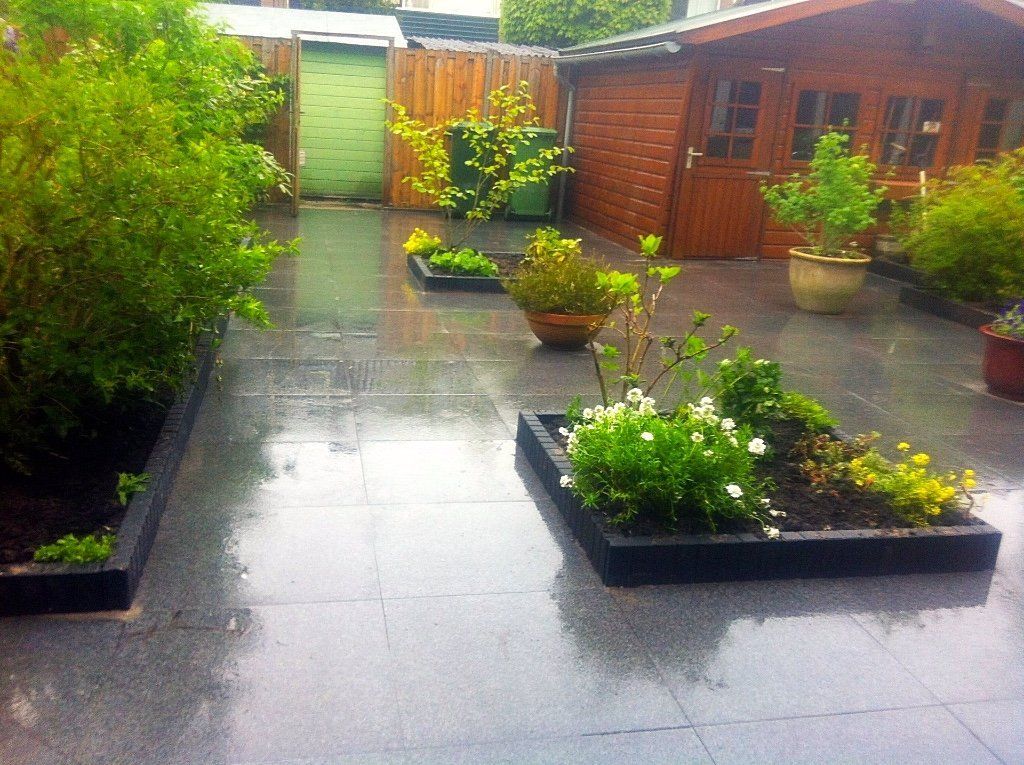 Een regenachtige achtertuin met grijze terrastegels, bloemperken, een houten schuur en een schutting.