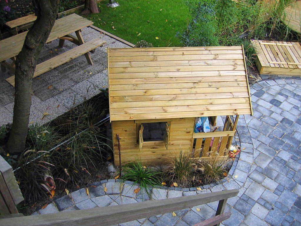 Houten speelhuisje met een schuin dak, op een stenen terras, vlakbij een picknicktafel en gras.