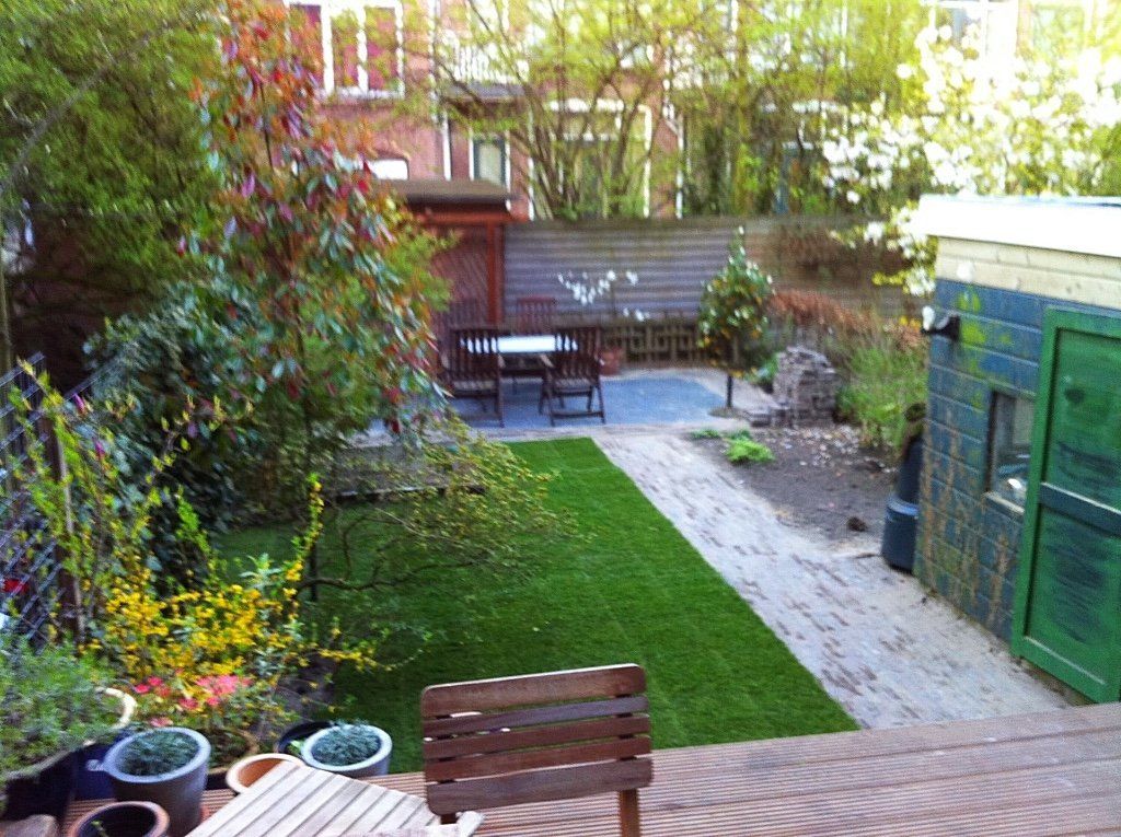 Achtertuin met groen gazon, stenen pad, houten terras, schuur en een tafel met stoelen.