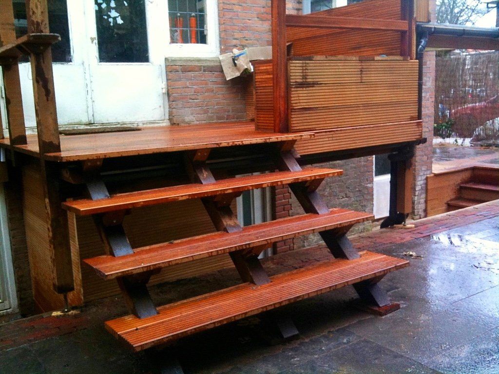 Een houten buitentrap met donkere steunbalken leidt naar een klein terras tegen een bakstenen gebouw.