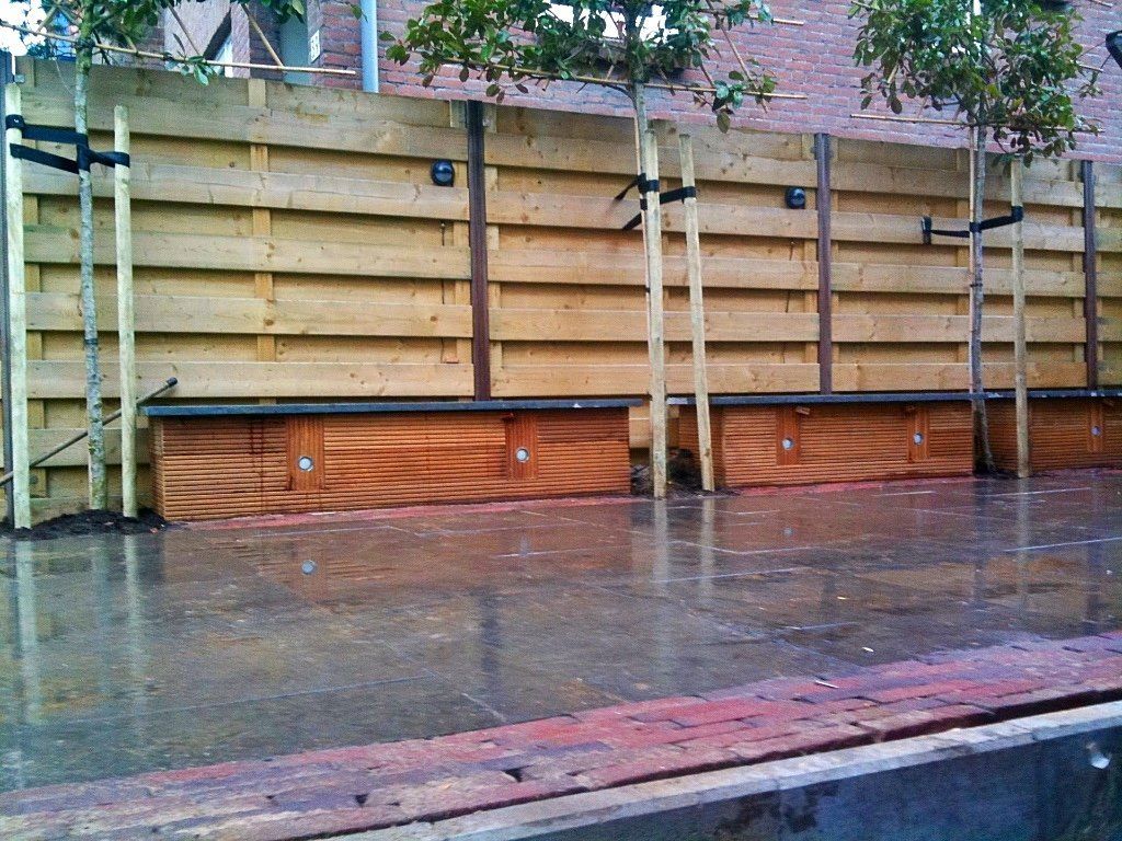 Houten schutting met daaraan vastgemaakte bomen, op een natte ondergrond van bakstenen en beton.