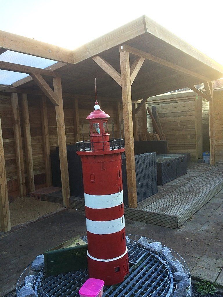 Een rood-wit vuurtorenbeeld staat op een rooster in een houten constructie in de tuin.