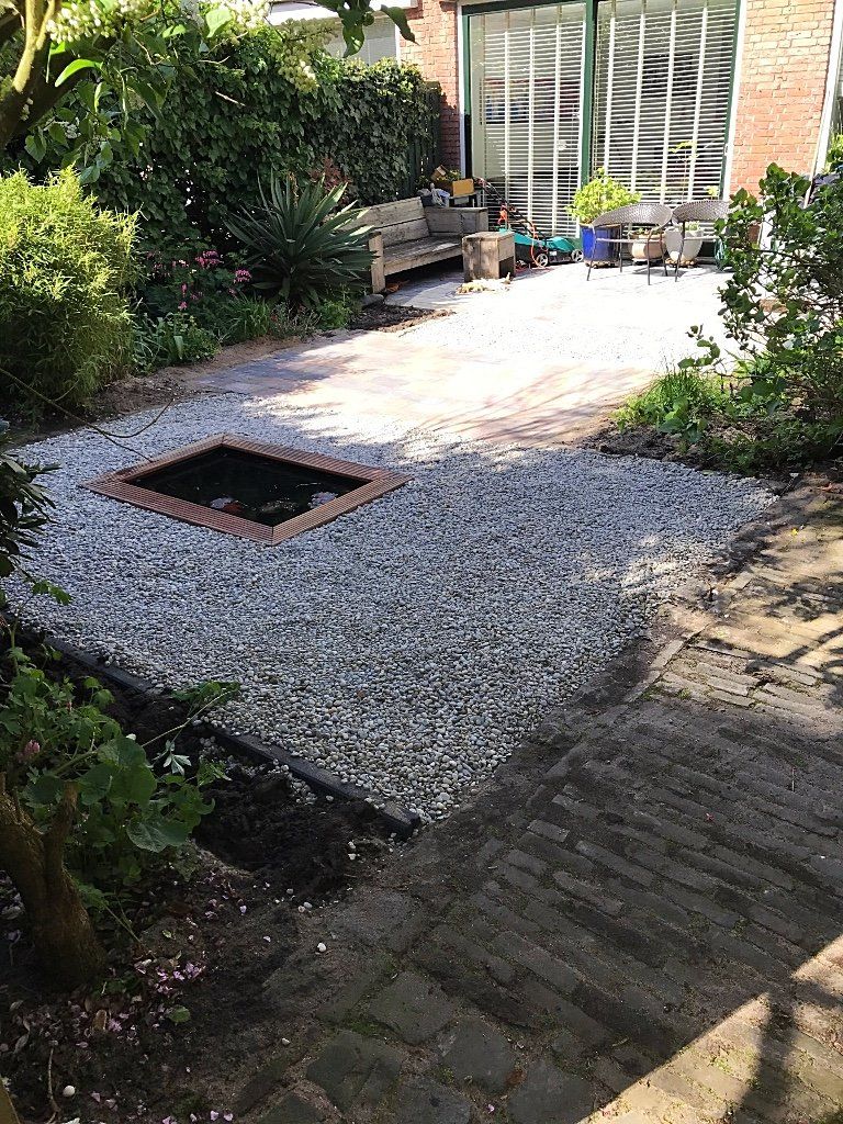 Terras in de achtertuin met grind, bakstenen, een vuurkuil, een bankje en groen.