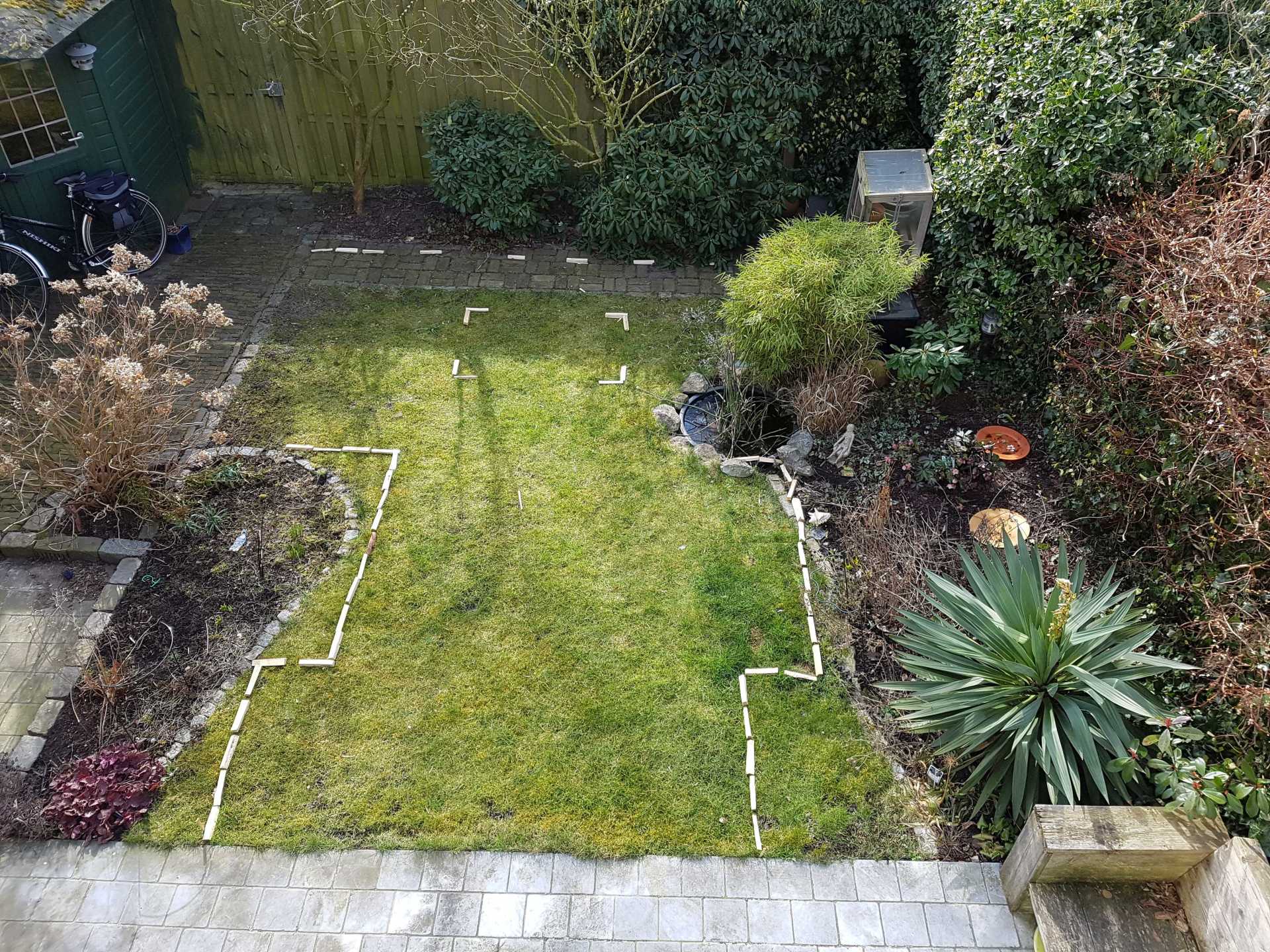 Bovenaanzicht van een achtertuin met een grasveld, omzoomd door bloembedden, struiken en een stenen terras.