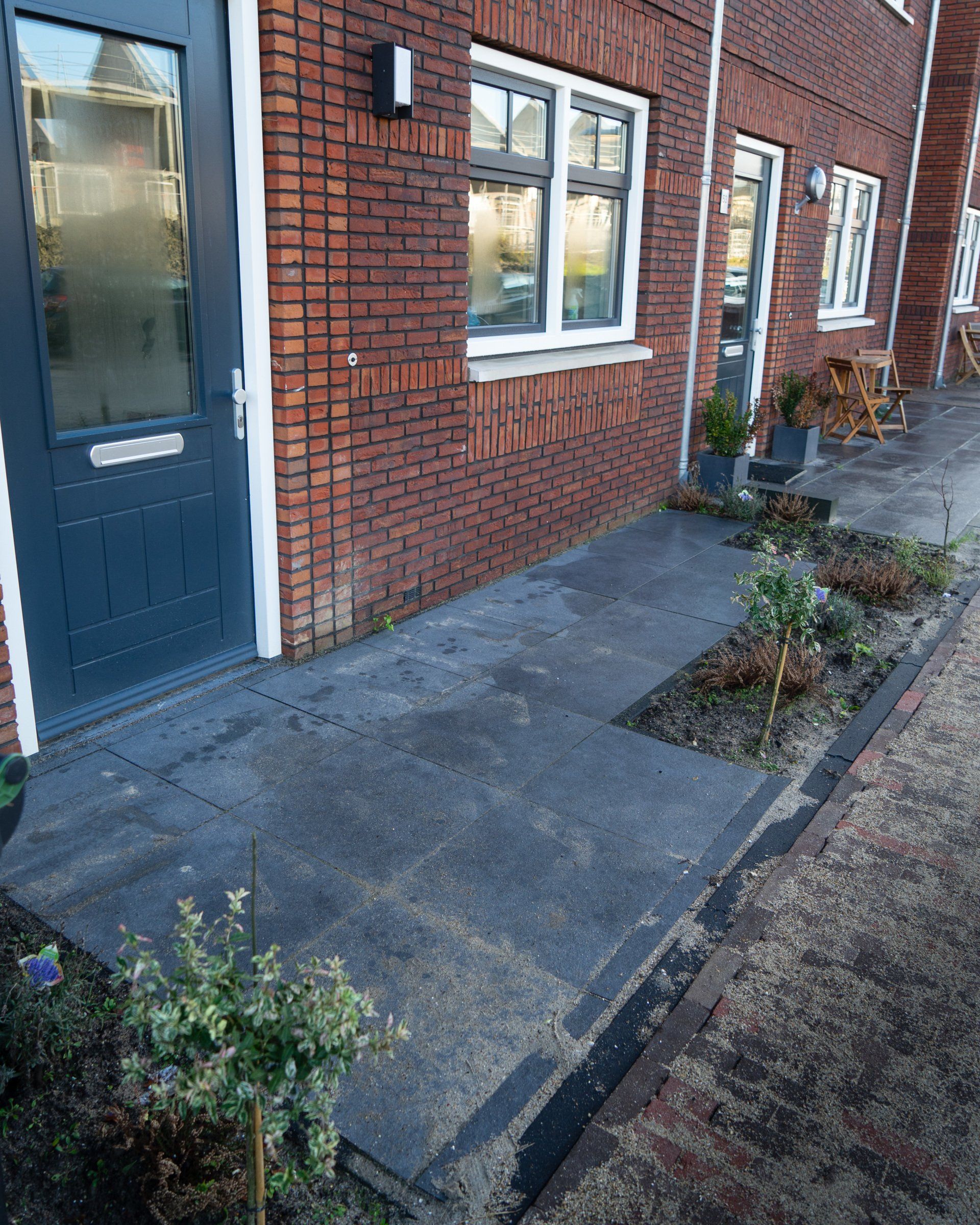 Een bakstenen gebouw met een donkerblauwe deur en een betonnen looppad. Langs het pad staan kleine plantjes.