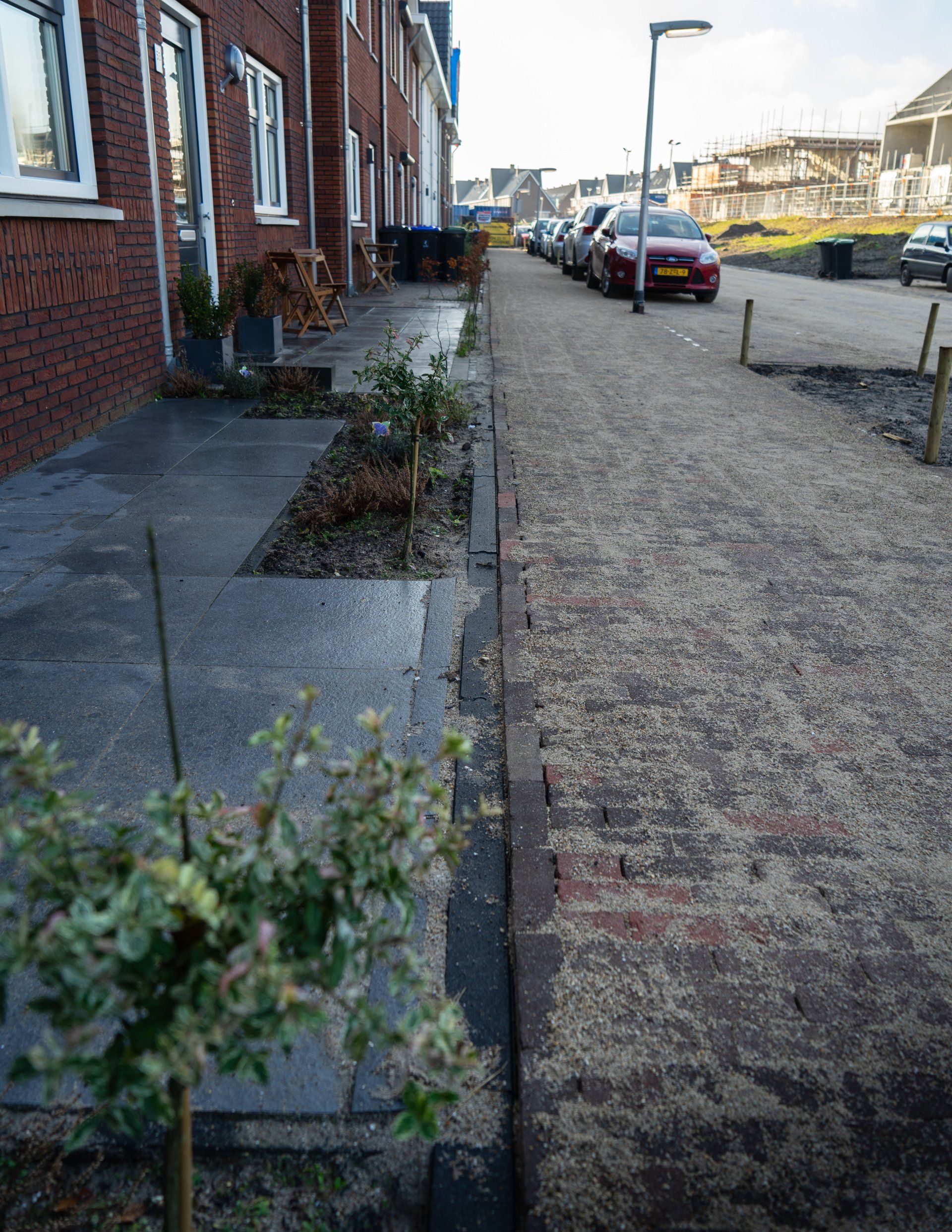 Een rij bakstenen rijtjeshuizen met een stoep, een grindpad en geparkeerde auto's op een zonnige dag.