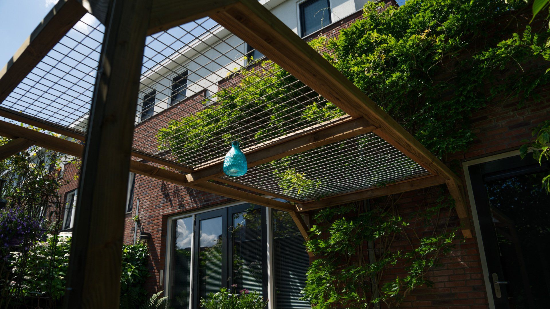 Houten pergola bedekt met gaas, begroeid met klimplanten. Een blauw object hangt aan de constructie, tegen de achtergrond van een bakstenen gebouw.