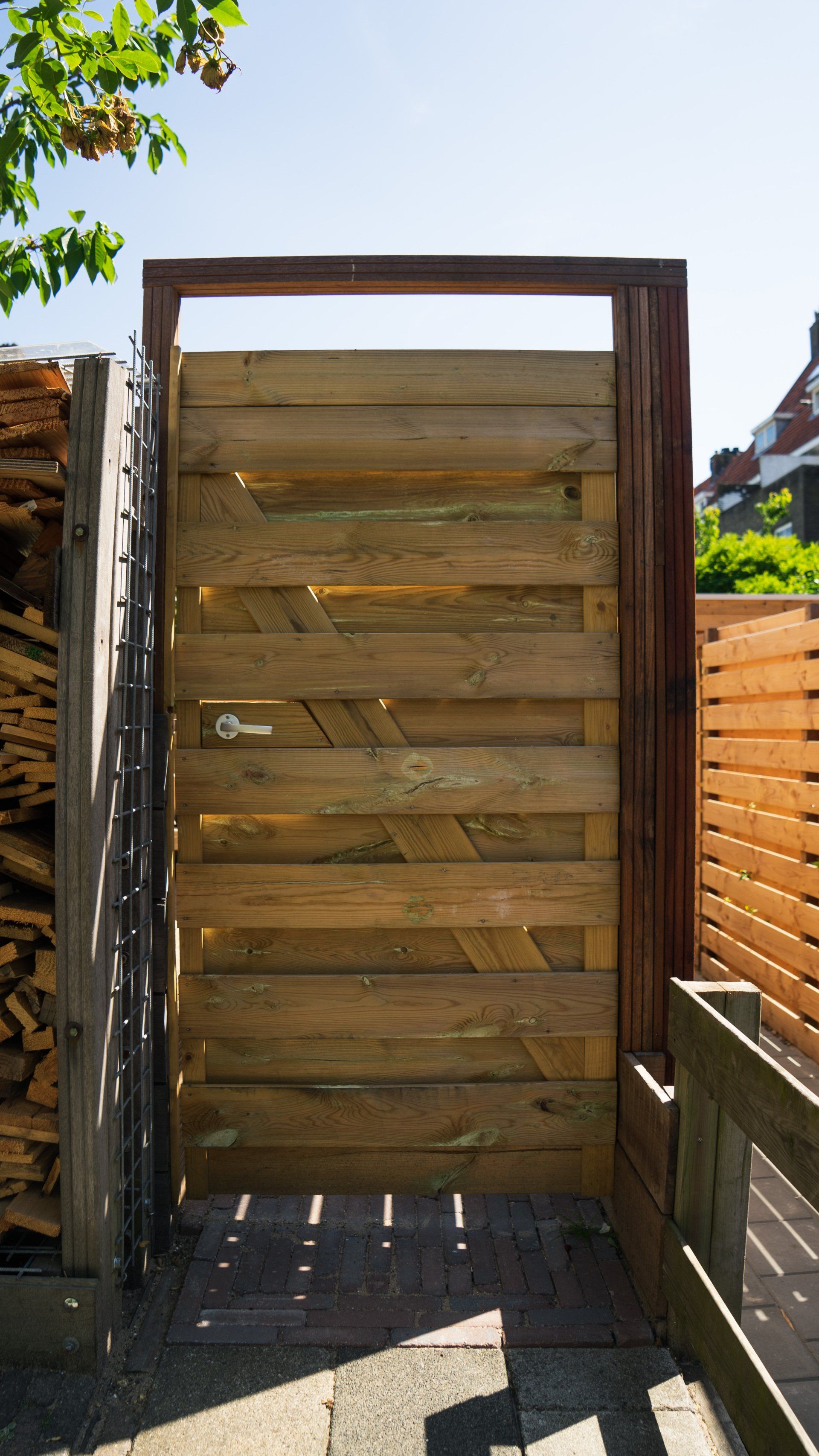Houten poort met horizontale planken, omlijst met bruin hout, die toegang geeft tot een zonnige buitenruimte.