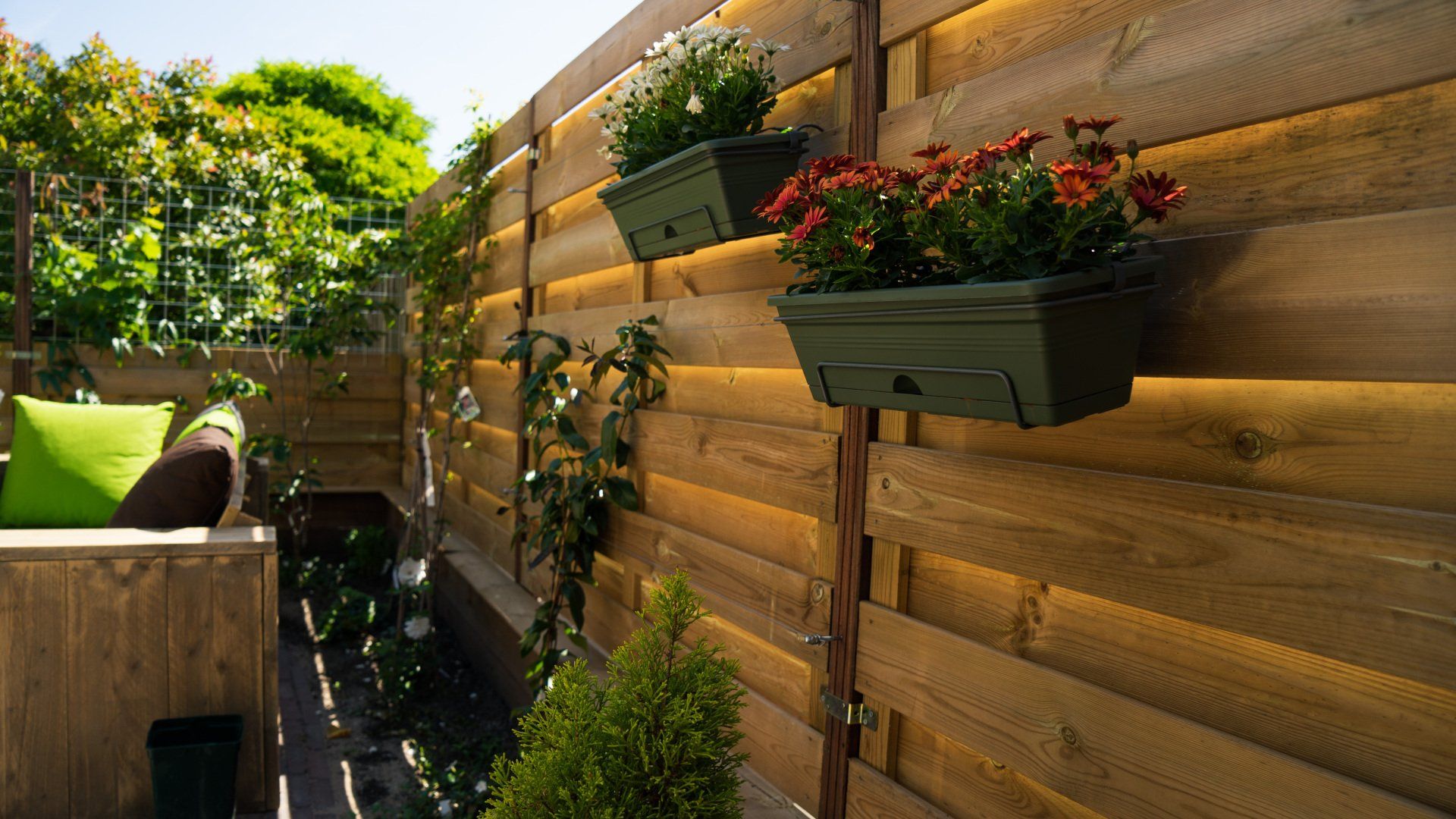 Houten schutting met hangende bloembakken; groene planten en een zitgedeelte met kussens.