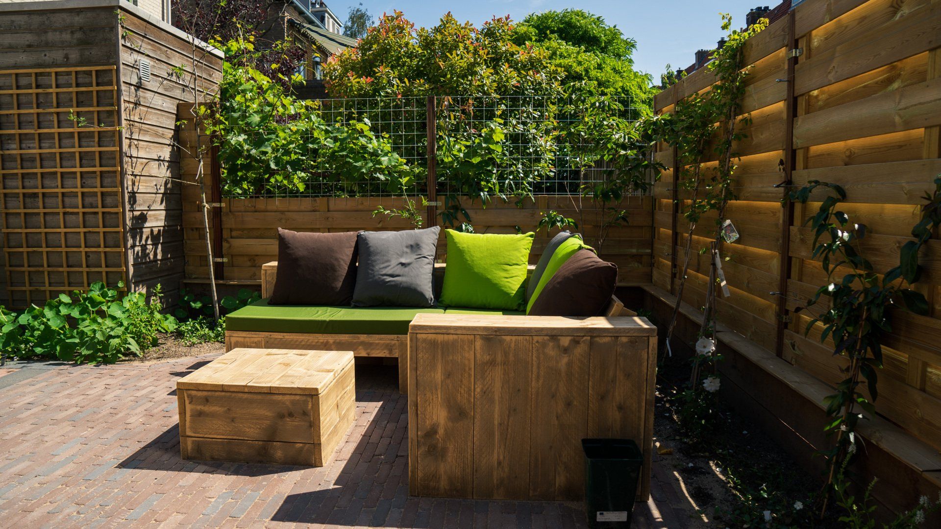 Terras met houten meubilair; bank met kussens, salontafel. Groene planten en een houten schutting op de achtergrond.