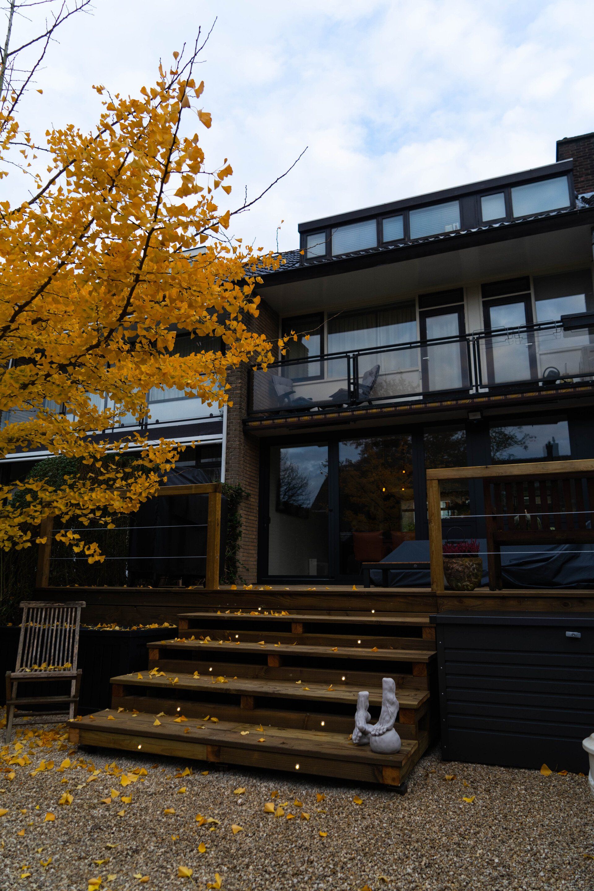 Achterkant van een modern huis met een houten terras en trap; gele herfstbladeren.
