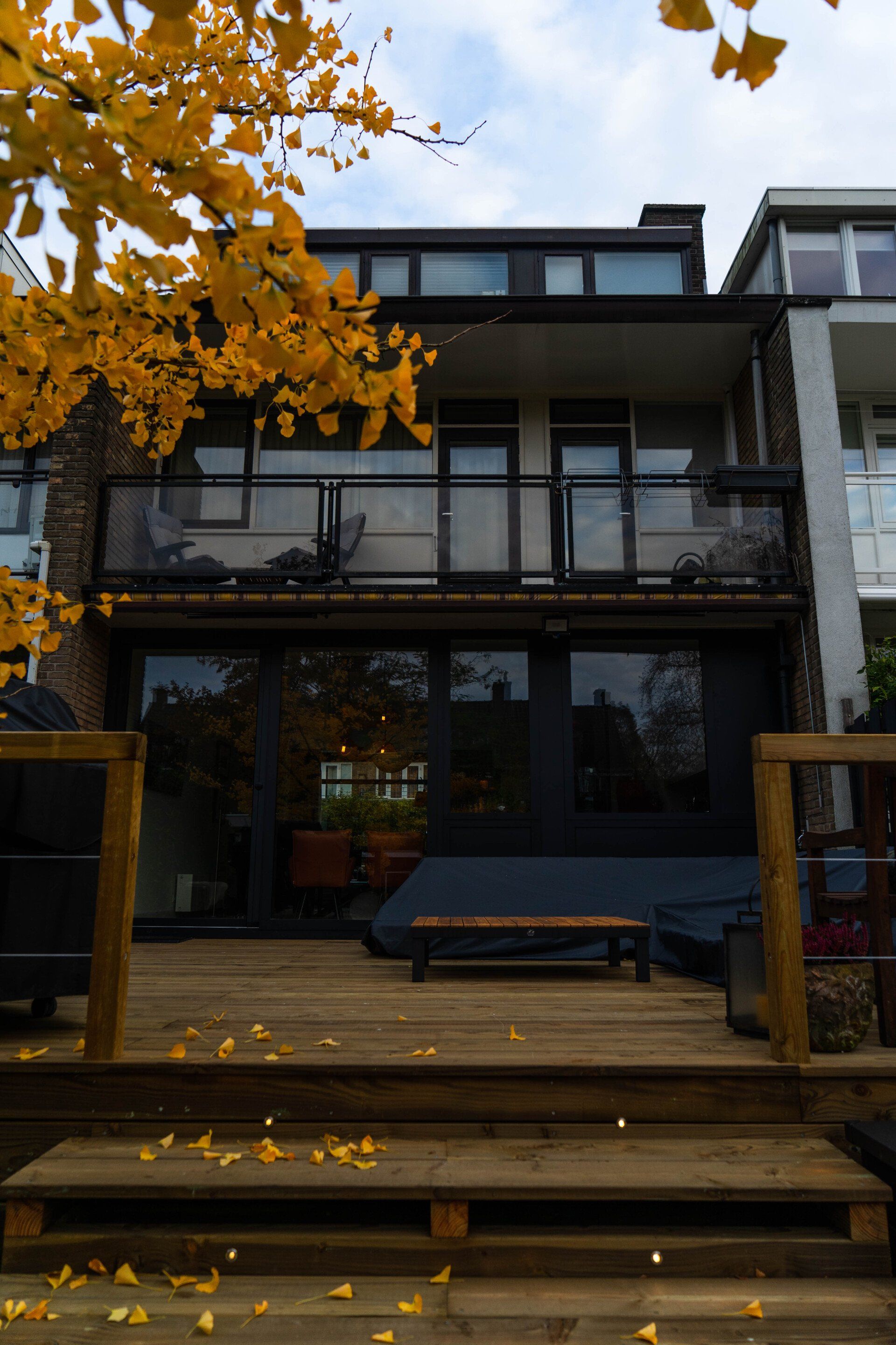 Een huis met meerdere verdiepingen, een houten terras, zwarte kozijnen en geel bladgroen.