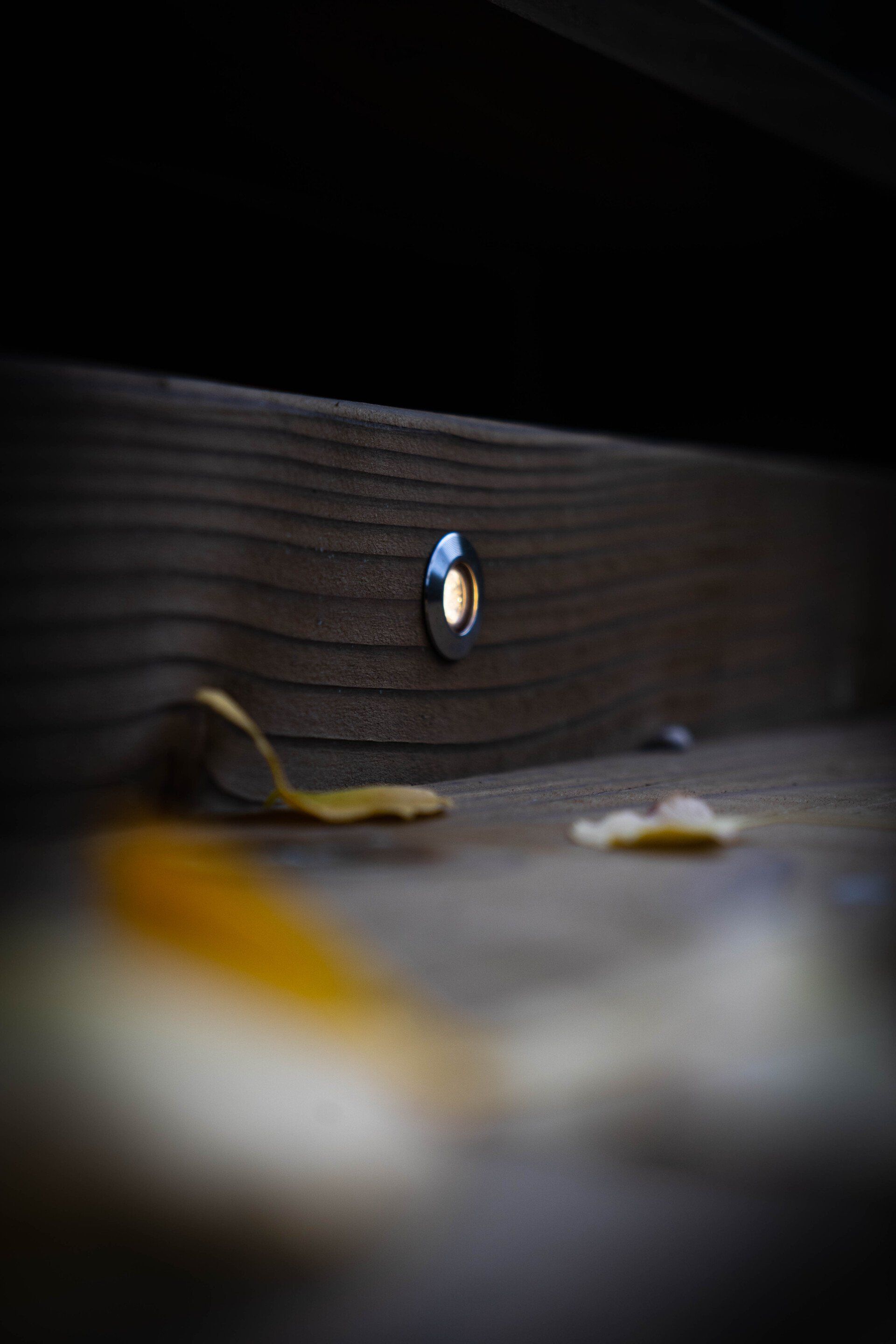 Een inbouwspot in een houten trede verlicht een klein gebiedje met herfstbladeren die er vlakbij verspreid liggen.