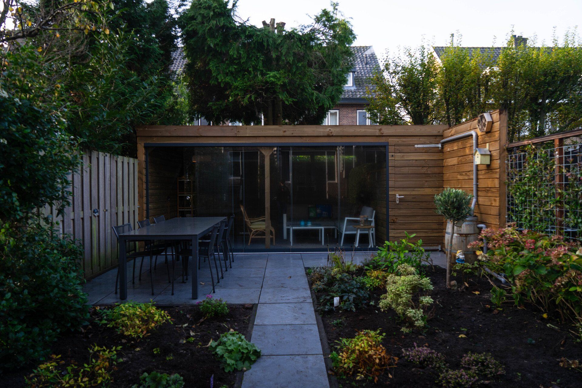 Houten tuinhuisje met glazen deuren, terras, tafel, stoelen en een weelderige tuin.