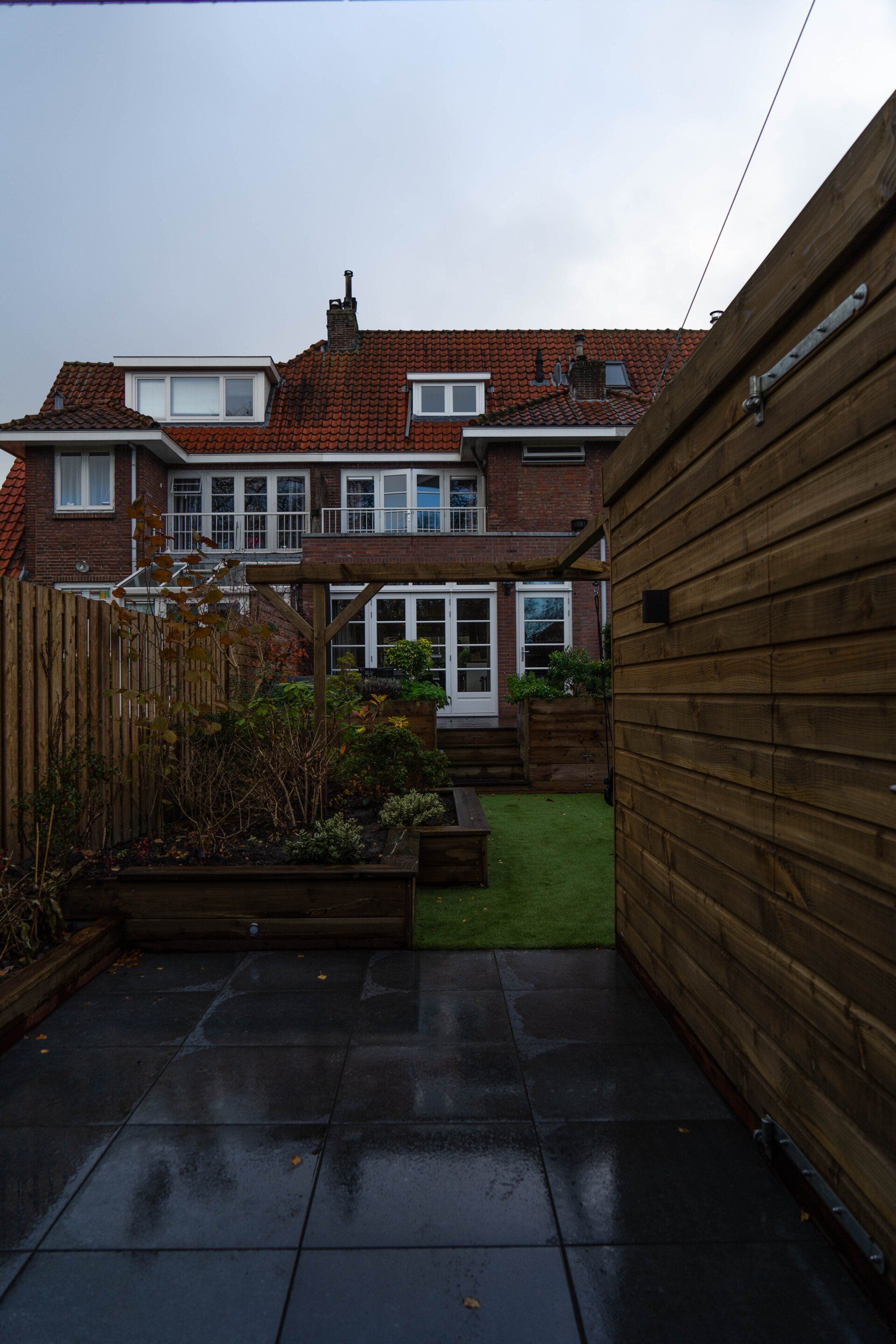 Uitzicht op de achtertuin van een bakstenen huis met een schuur op een natte, grijze dag. Tuin met plantenbakken en gras.