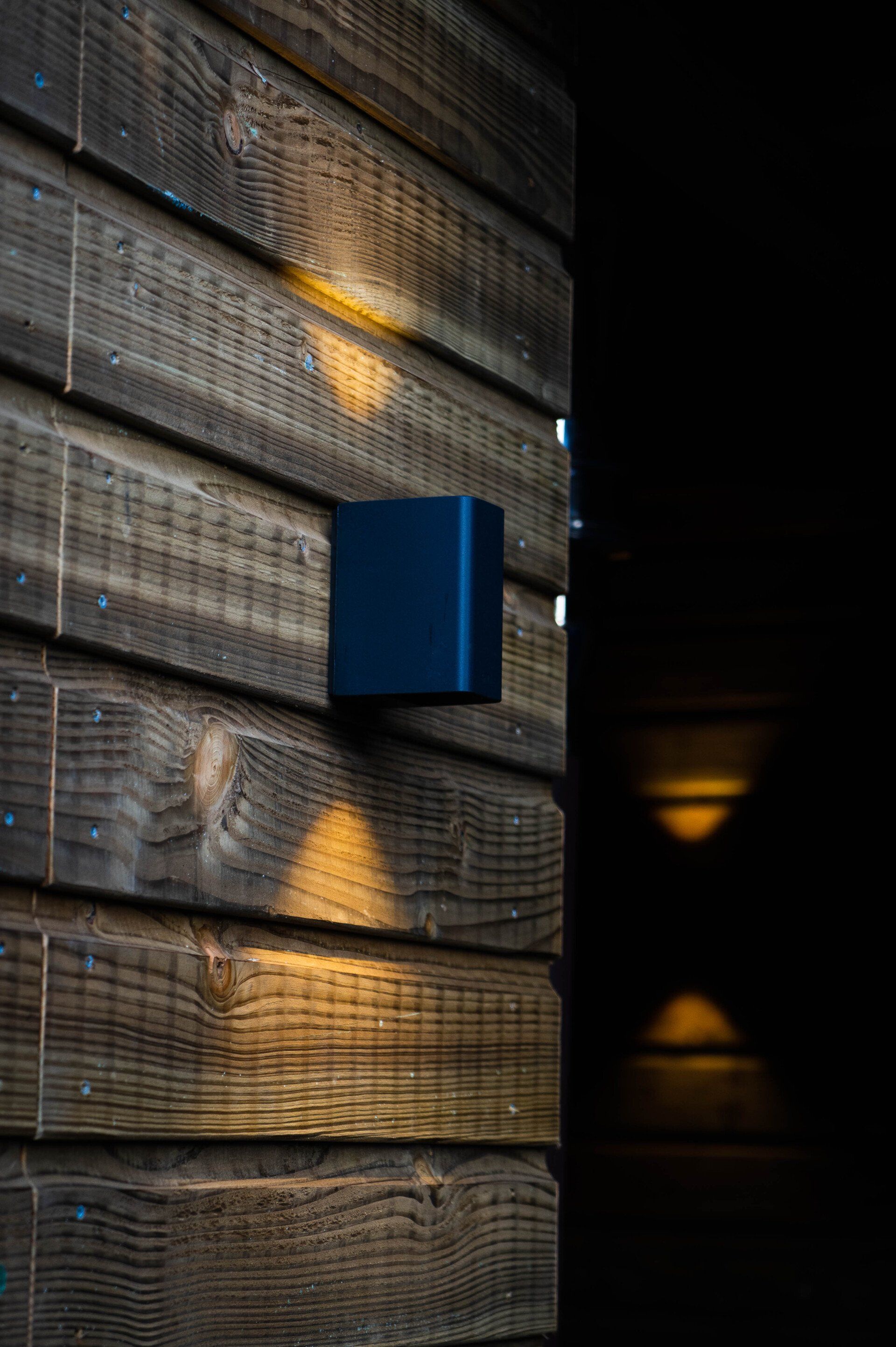 Zwarte wandlamp op een houten gevelbekleding met structuur, die driehoekige lichtpatronen projecteert.