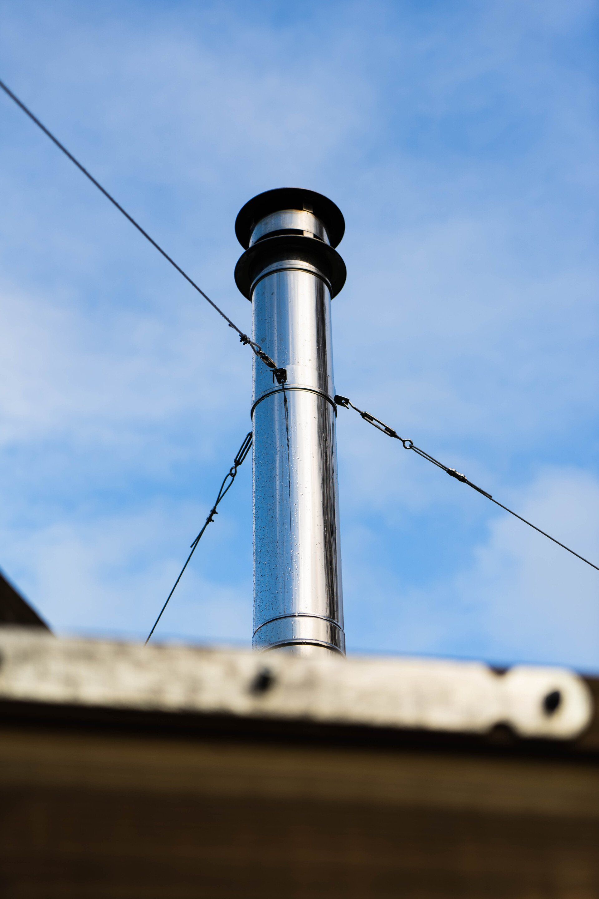 Een glanzende metalen schoorsteen tegen een blauwe lucht, ondersteund door draden, op een dak.