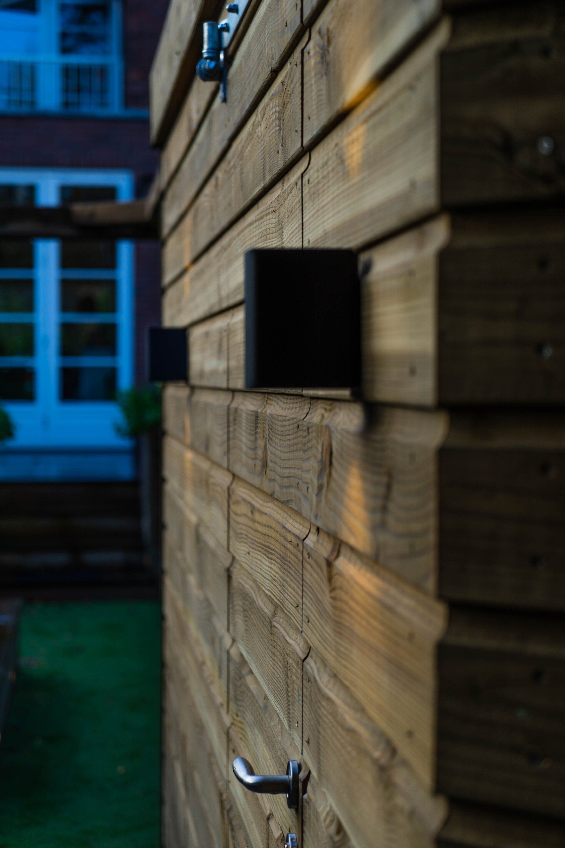 Houten gebouw met zwarte vierkante lampen en zilveren handgreep, gras en een blauw raam op de achtergrond.