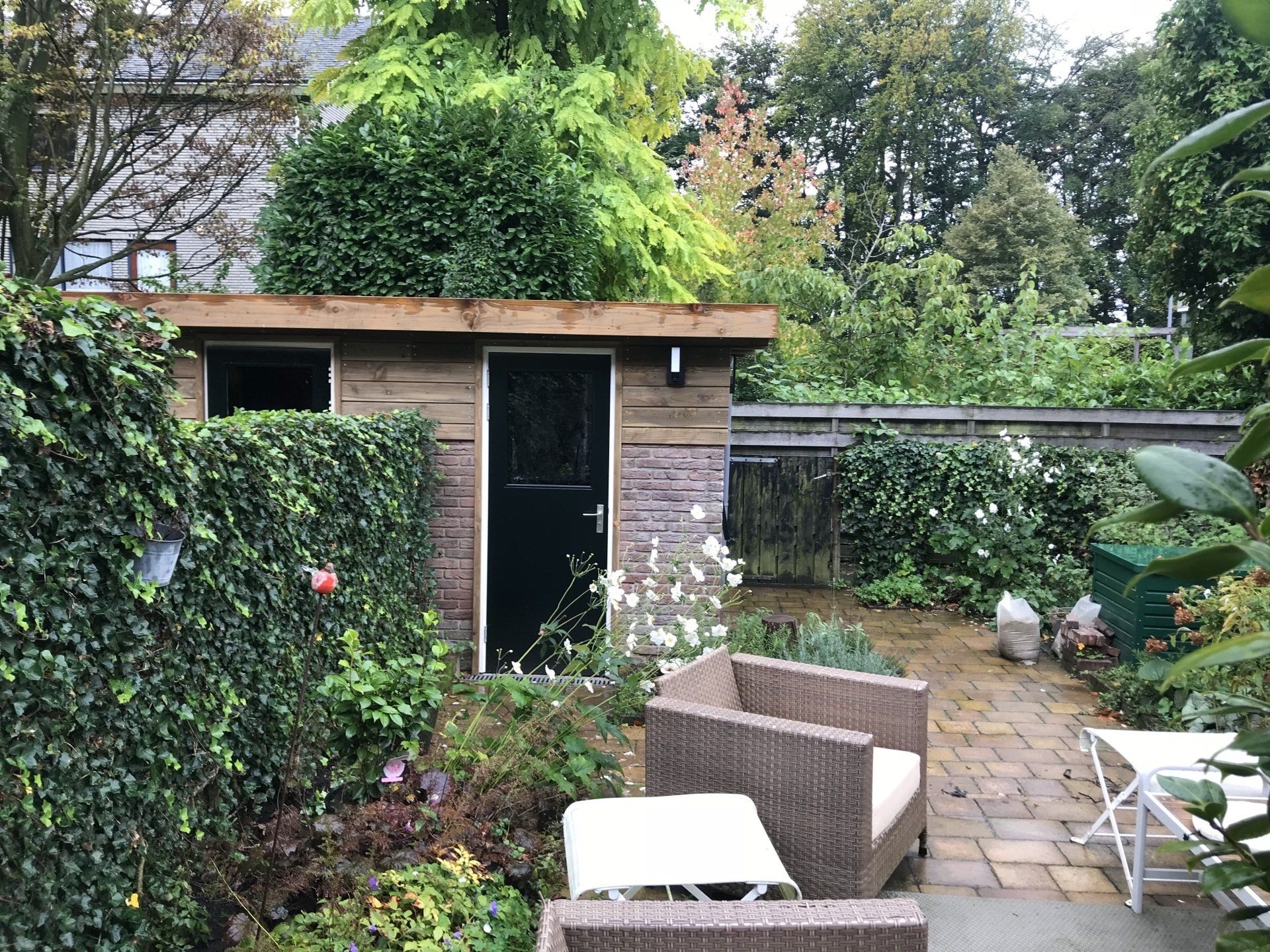 Achtertuin met schuur, twee rieten stoelen, een kleine tafel en weelderig groen.