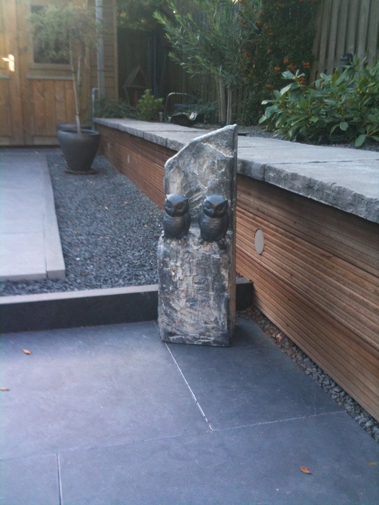 Stenen sculptuur van twee uilen in een tuin met een houten keermuur en terras.