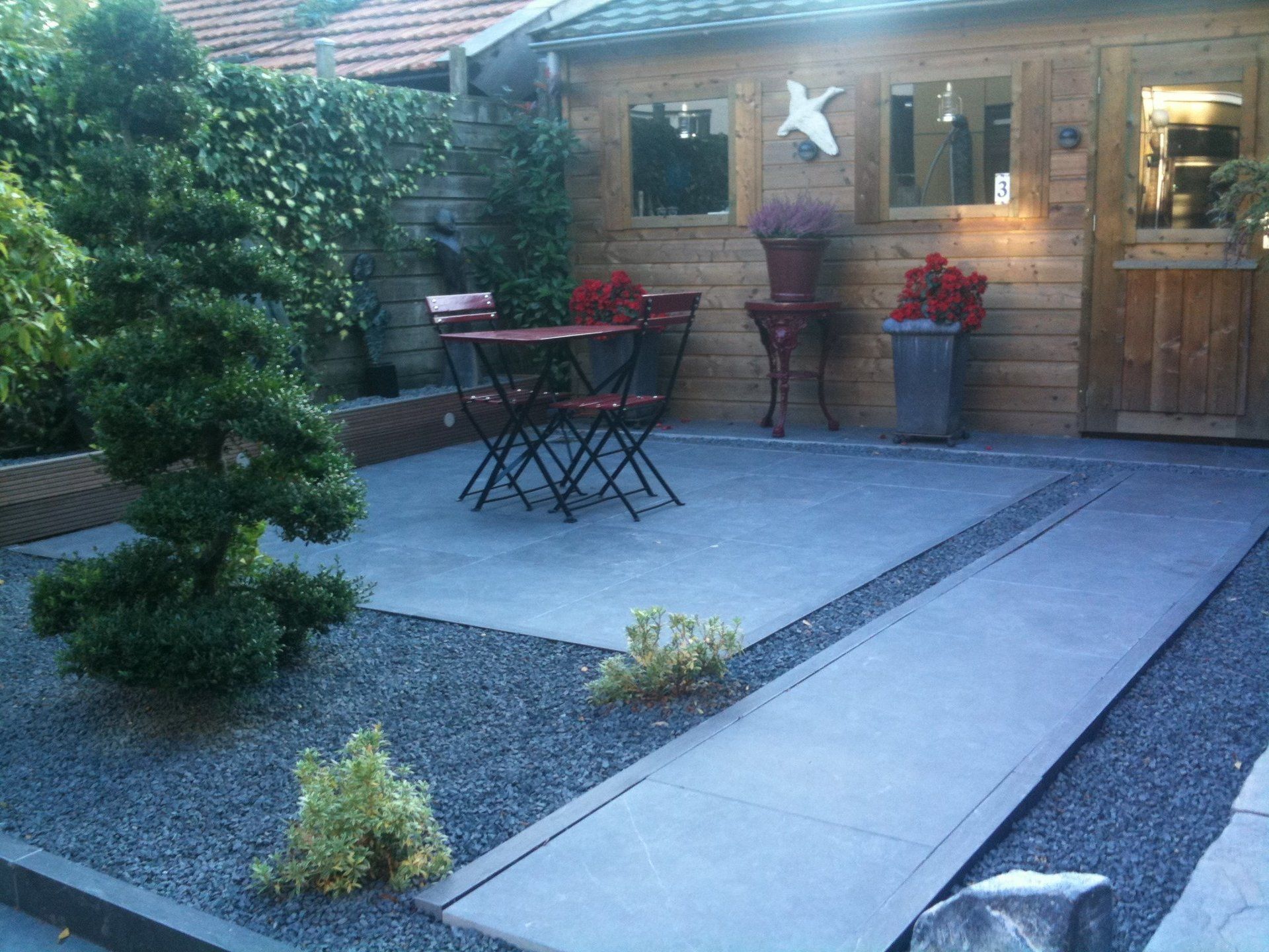 Terras buiten met grind, betonnen looppad, rode tafel en stoelen, houten schuur.