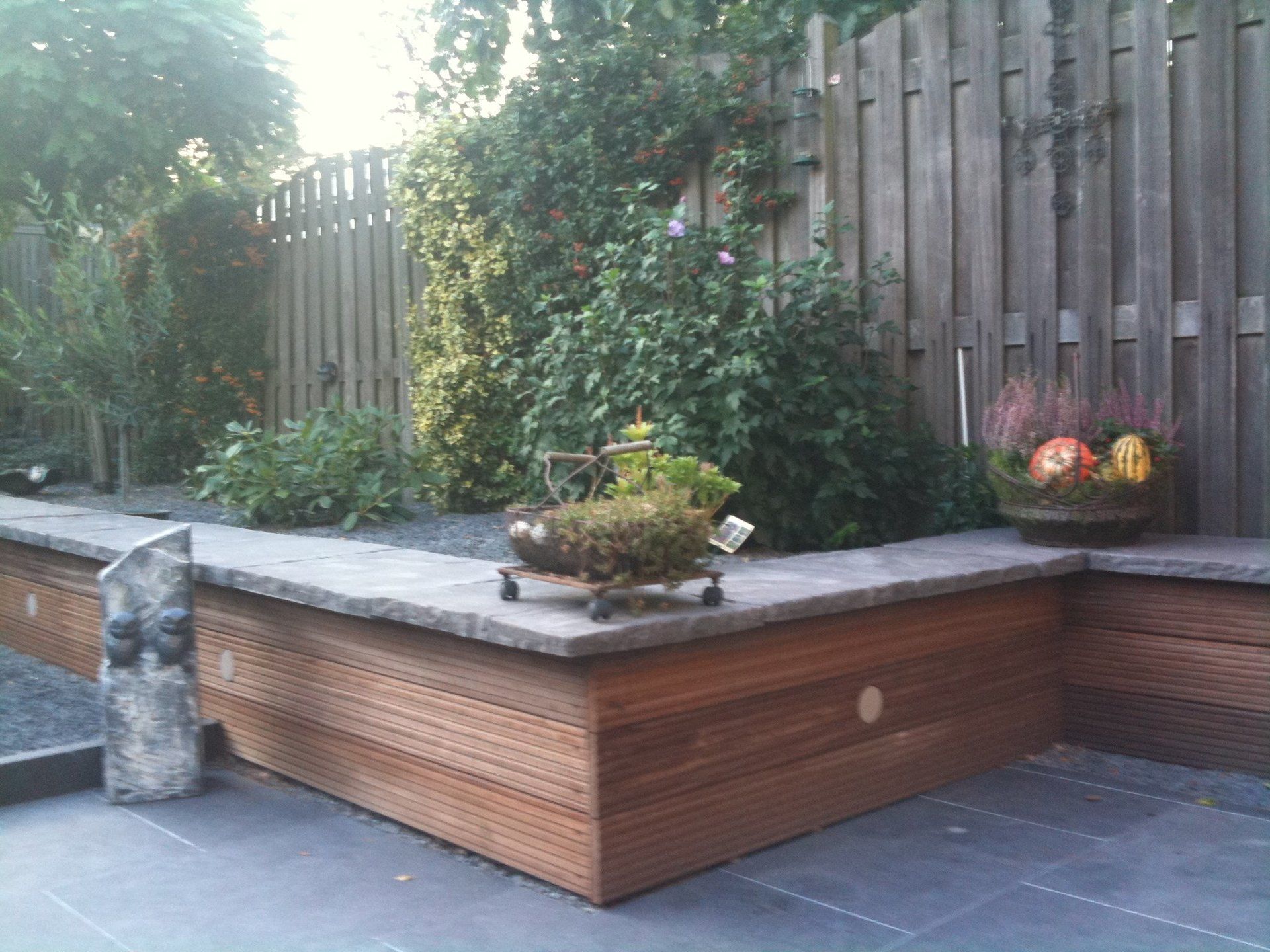 Houten bloembedden met planten en een decoratieve kar, geplaatst tegen een houten schutting en groen.
