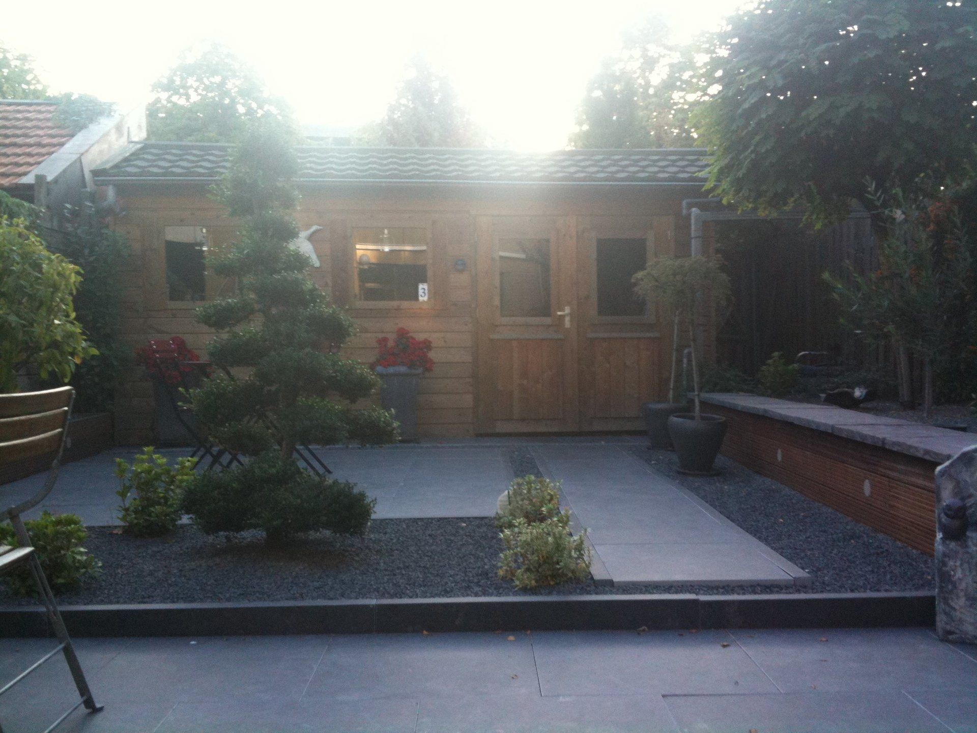 Houten schuur in een achtertuin met een stenen terras, een bonsaiboom en grindborders.