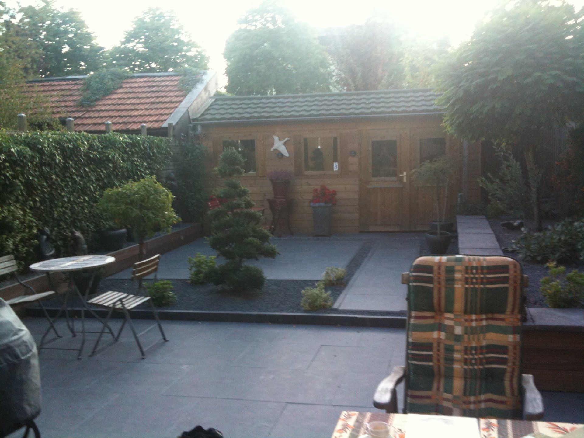 Terras in de achtertuin met houten schuur, bomen, zitgelegenheid en een bestraat gedeelte.