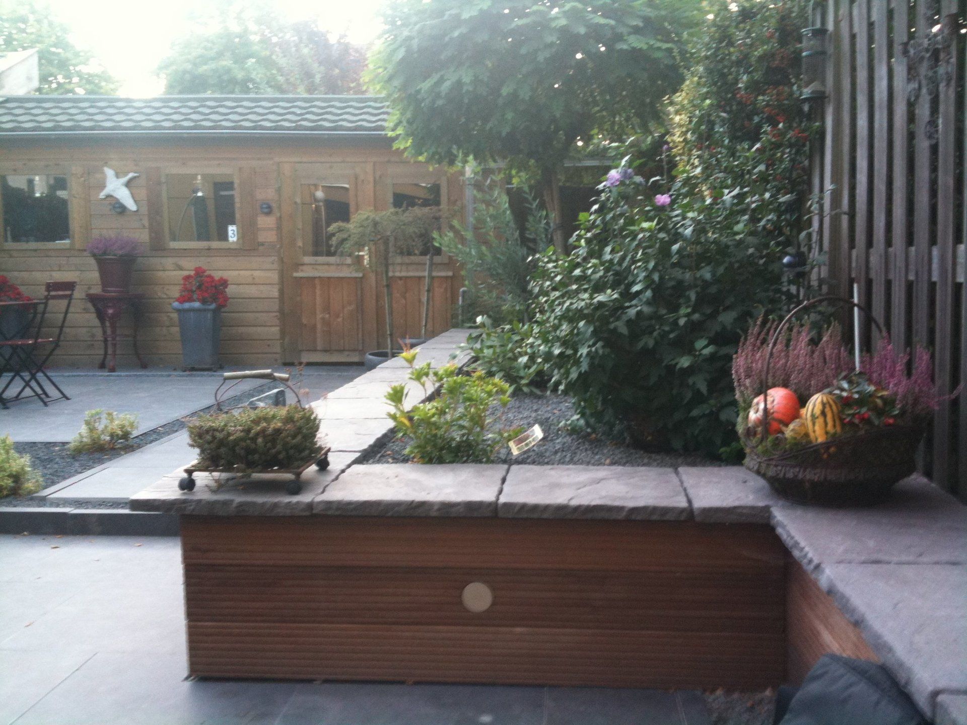 Houten schuur en terras met ingebouwde bank, omgeven door planten en een houten schutting.