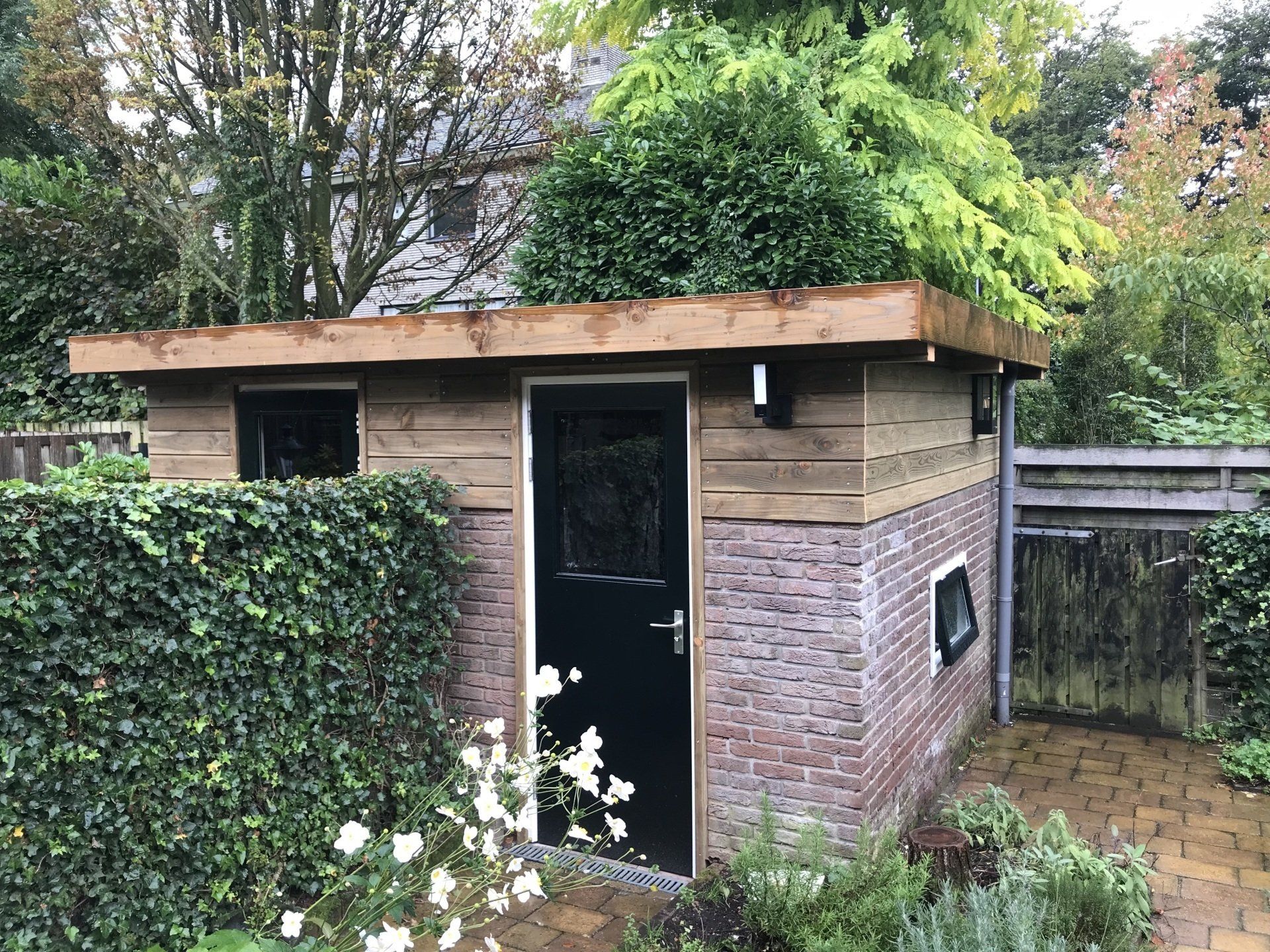Een klein schuurtje van baksteen en hout met een donkere deur, ramen en een groen dak, omgeven door groen.