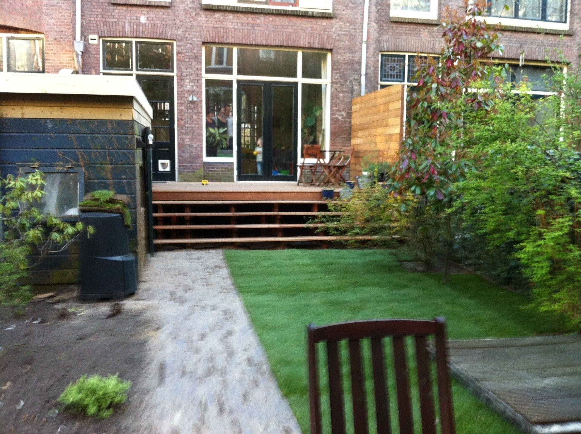 Achtertuin met houten terras, trap en groen gazon, die leidt naar een bakstenen gebouw met glazen deuren en ramen.