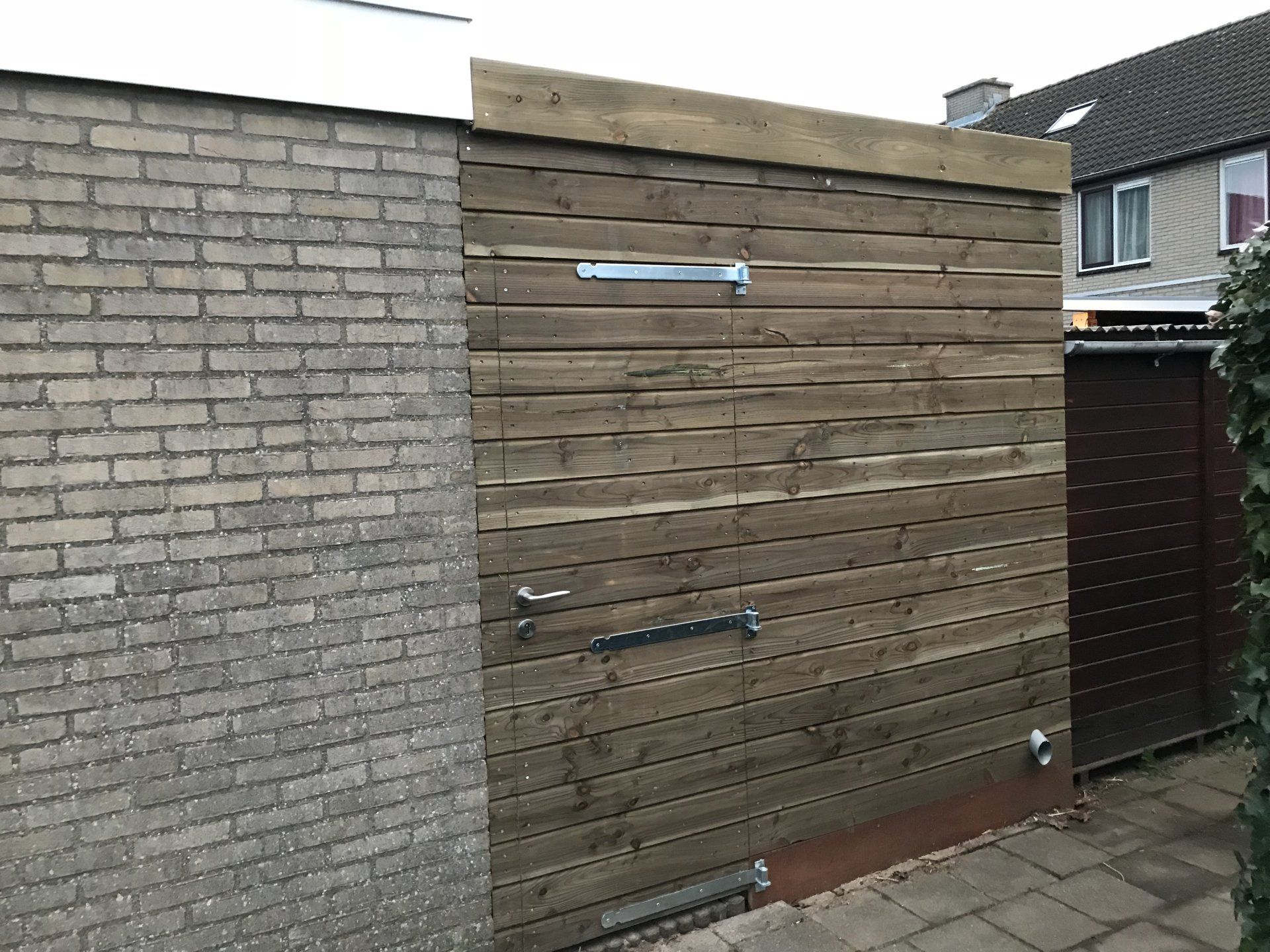 Houten poort en bakstenen muur op een bestraat oppervlak, bruin hout, grijze bakstenen en een huis op de achtergrond.