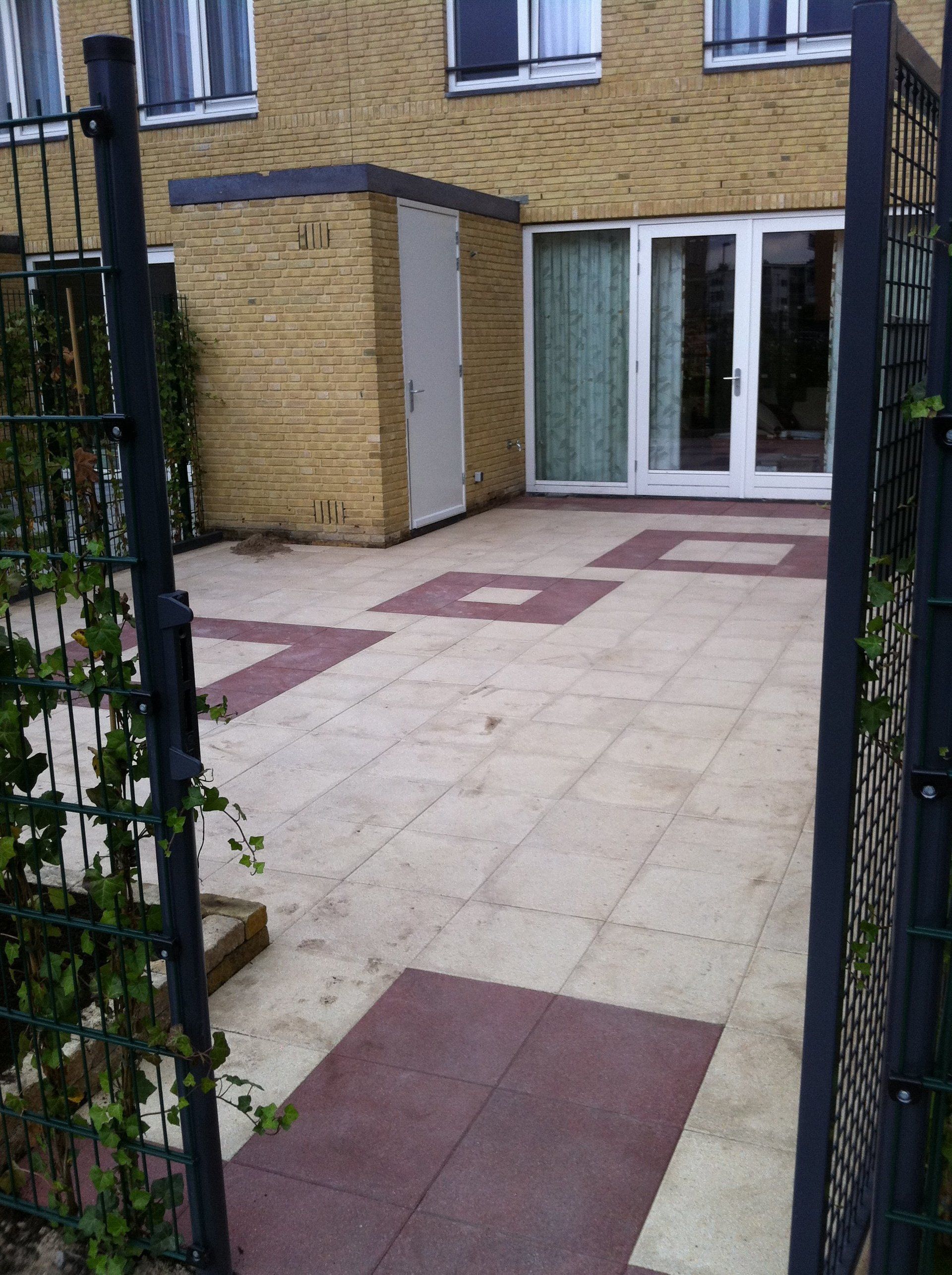 Terras met beige en rode tegels, schuur, glazen schuifdeur en zwart metalen hek.